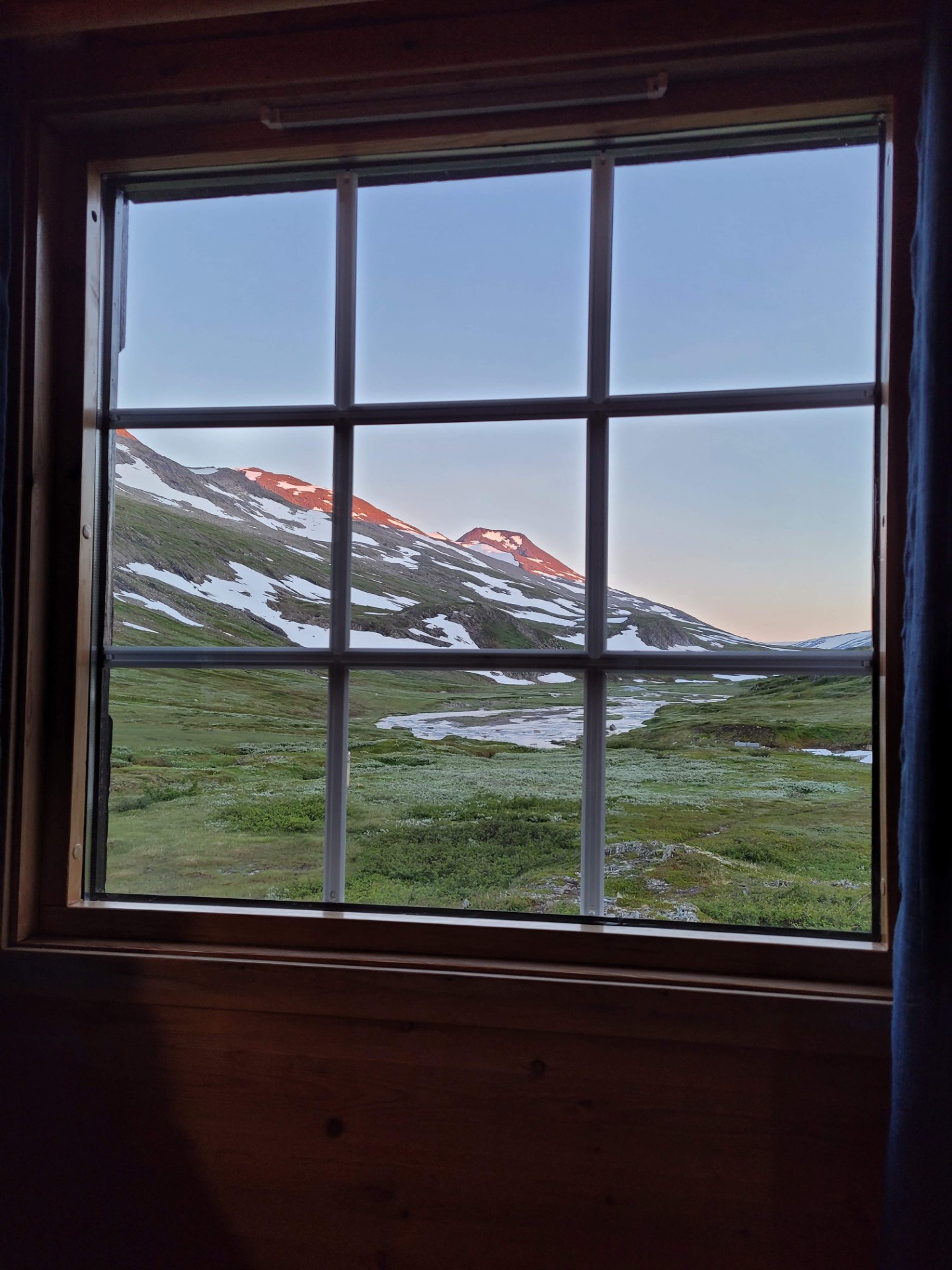 Utsikt fra et hyttevindu. Man ser ut på et høyfjellandskap med gress, vann og fjelltopper men noen snøflekker på.